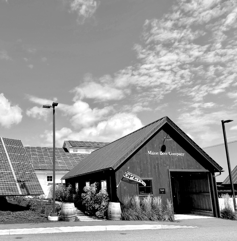 A black and white photo of the Black Barn entrance to Maine Beer Company's tasting room with a large solar tracker off to the left.