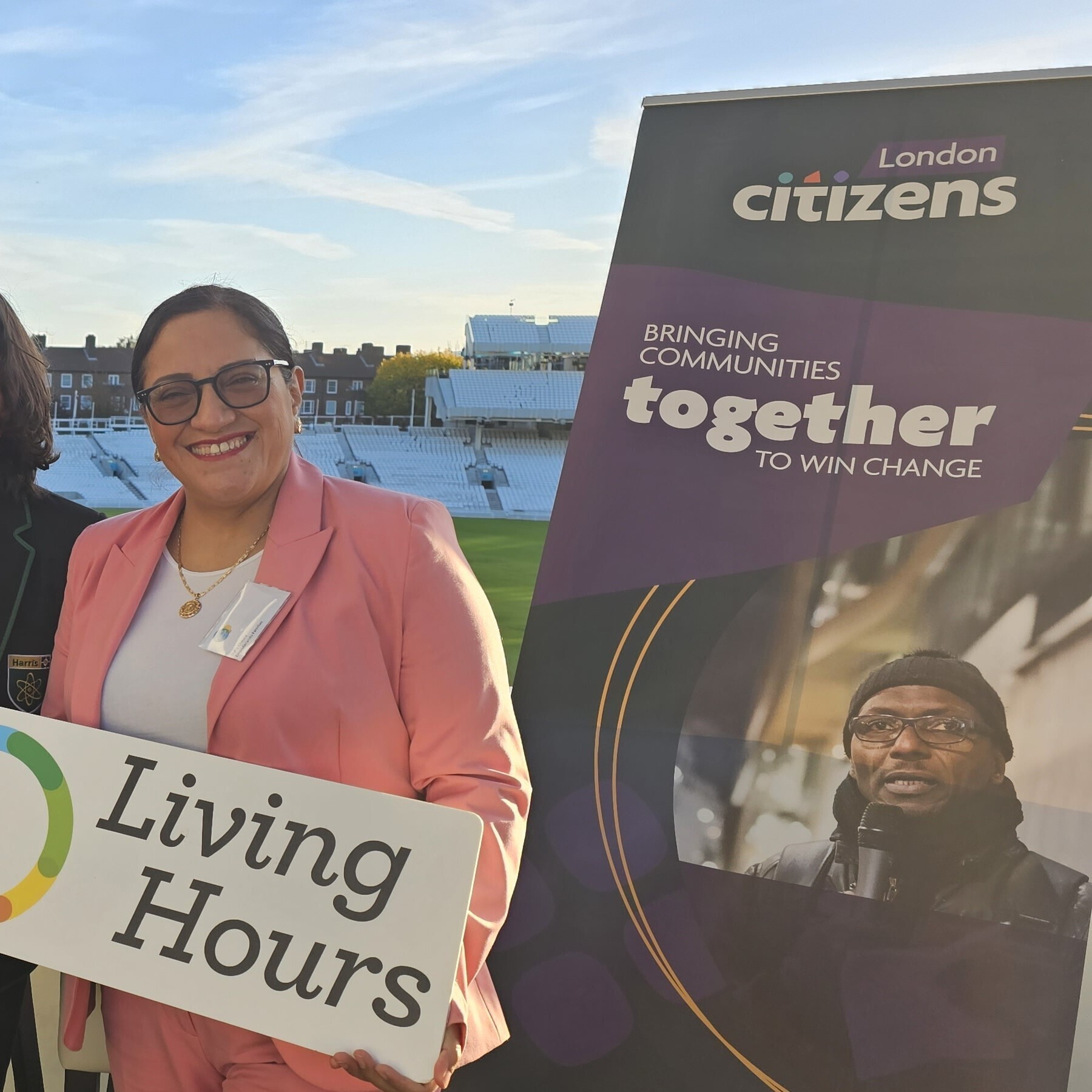 Doris is holding a sign that reads 'Living Hours' and is front of a Citizens UK sign that reads 'Bringing communities together to win change'. She is smiling at the camera, wearing a pink suit.