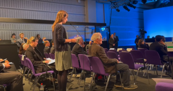 ShareAction Head of Banking Programme, Jeanne Martin, is shown at a Barclays' AGM asking a question. She is stood up with notes in her hand, speaking into a microphone and surrounded by other attendees sat in chairs.