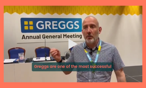 A screenshot of a video diary of our day at the Greggs AGM. The screenshot shows a man in front of a big sign that says 'Greggs Annual General Meeting' and a long table with chairs behind it. The man is mid-sentence, using his hands to express what he is saying. He is wearing a shirt and a lanyard. He is captioned to be saying
